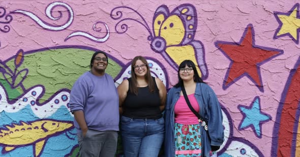 Three individuals stand in front of large mural painted on the outside wall of a building.